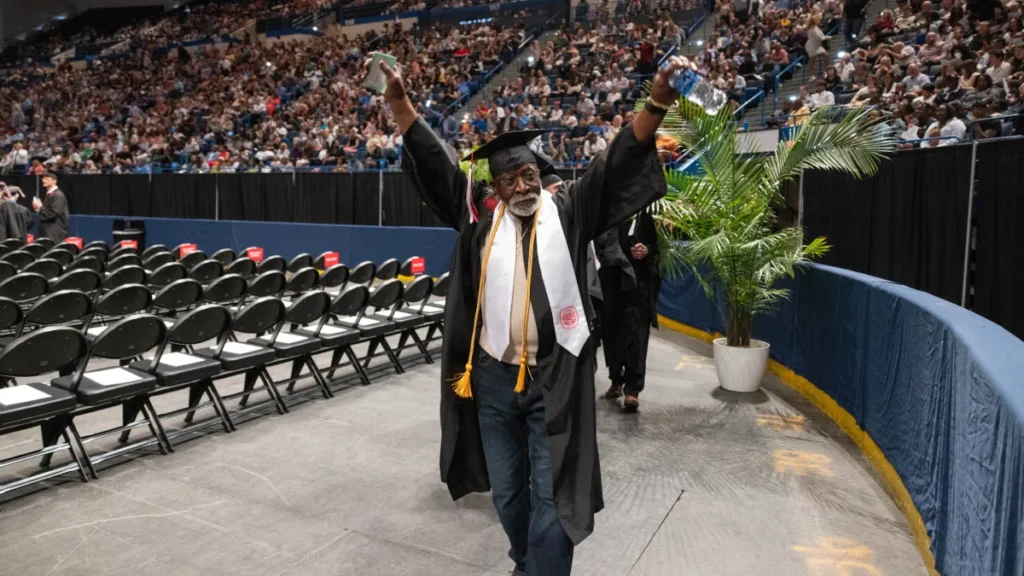 A man in cap and gown walking across the floor of an arena.