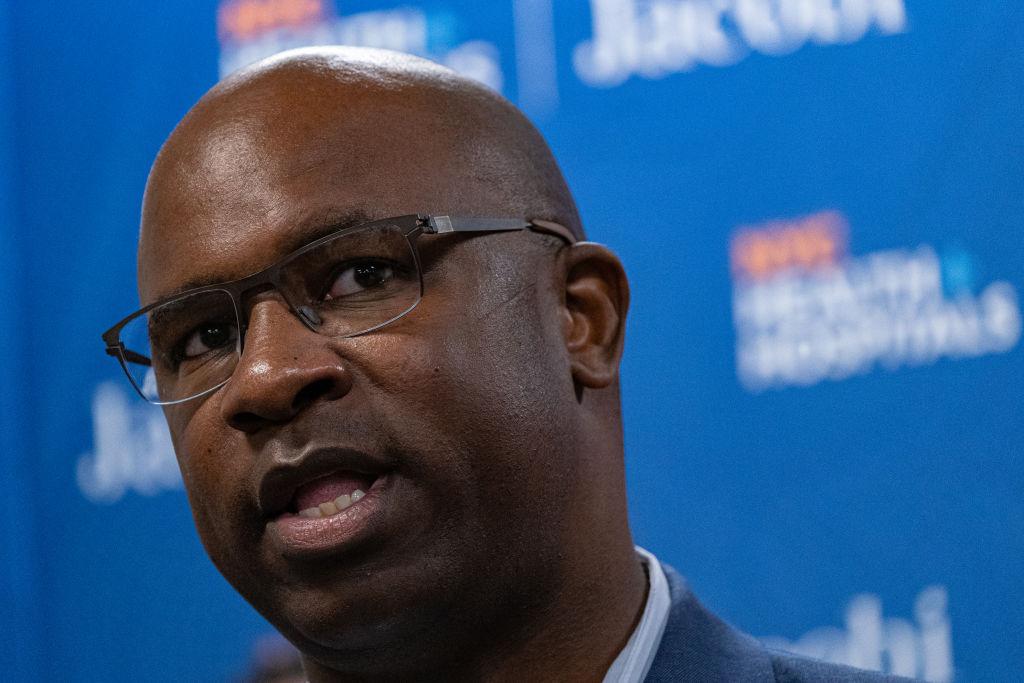 A man with glasses is standing in front of a blue wall.